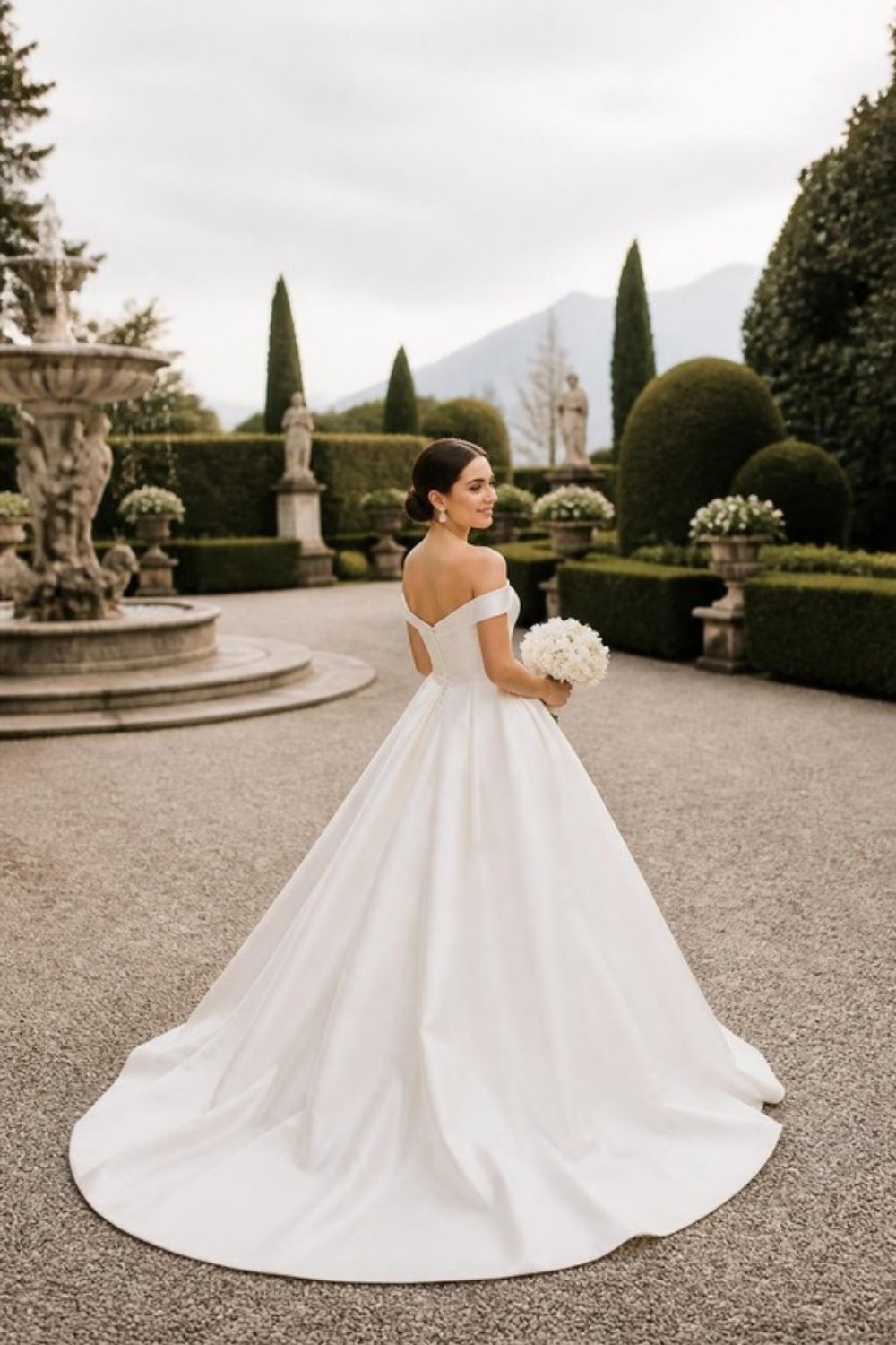 Bride in an off-the-shoulder ivory satin ball gown with a sleek low bun, holding white flowers in a formal garden
