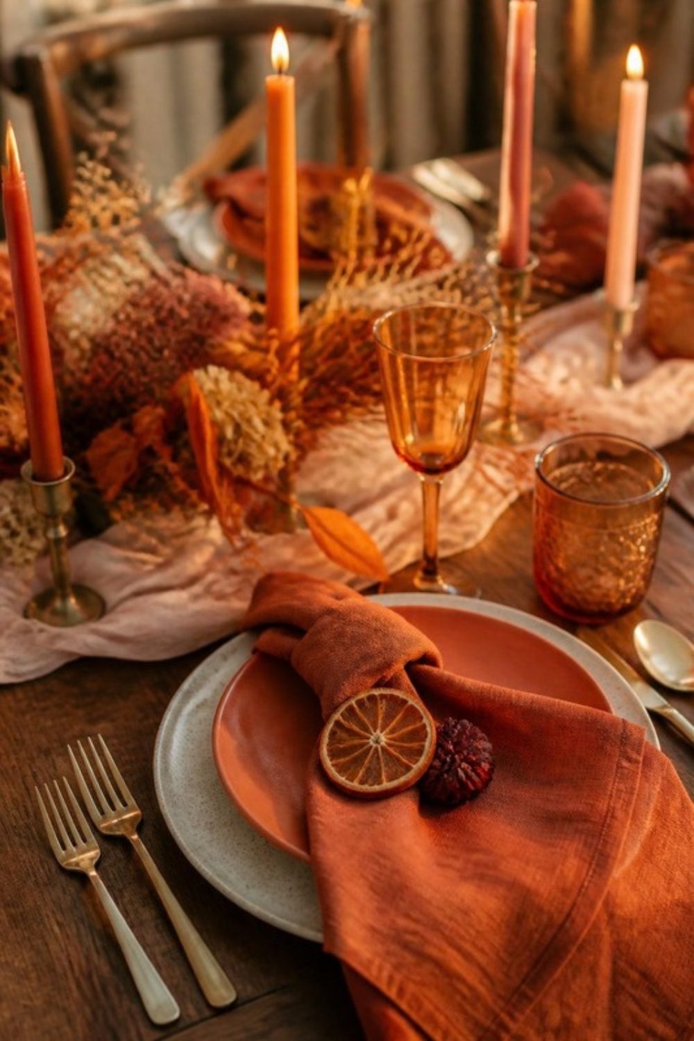 Terracotta place setting with a rust linen napkin, a dried orange slice, an amber glass and lit taper candles