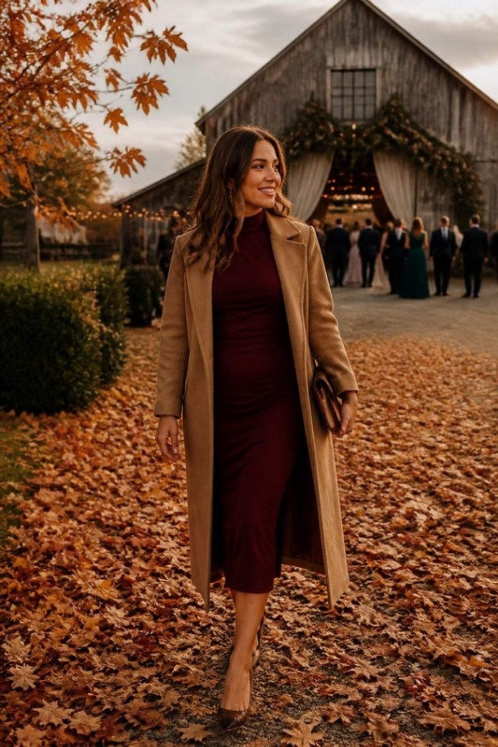 Guest in a burgundy long-sleeve midi dress and camel coat walking through fallen leaves toward a barn wedding