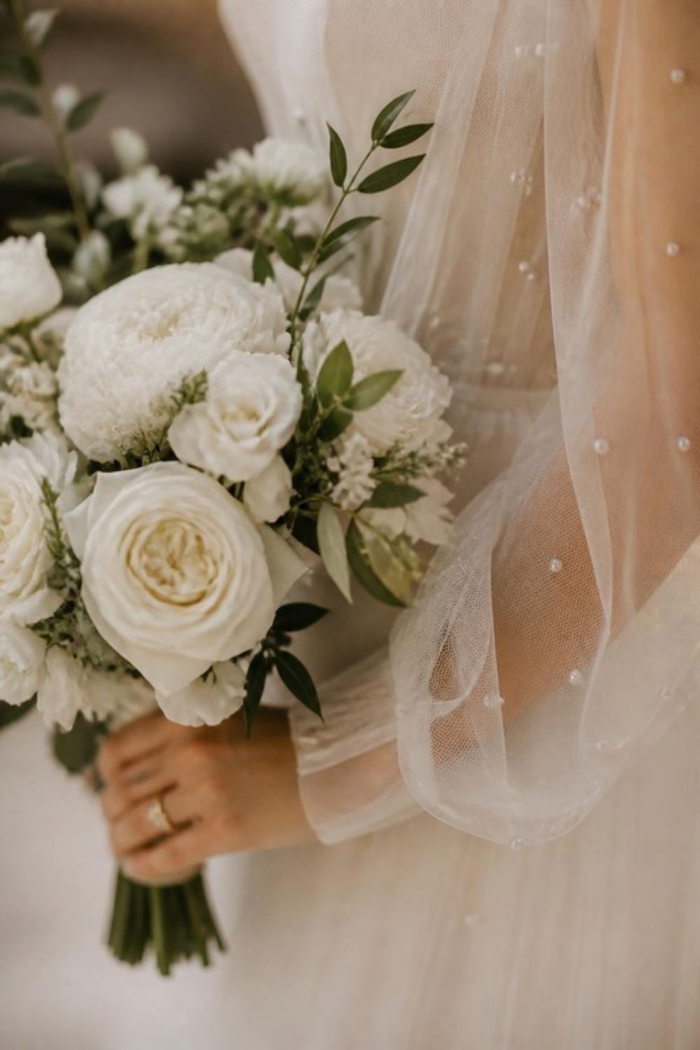 Detail of a sheer tulle wedding dress sleeve dotted with tiny pearls as the bride holds a white garden rose bouquet